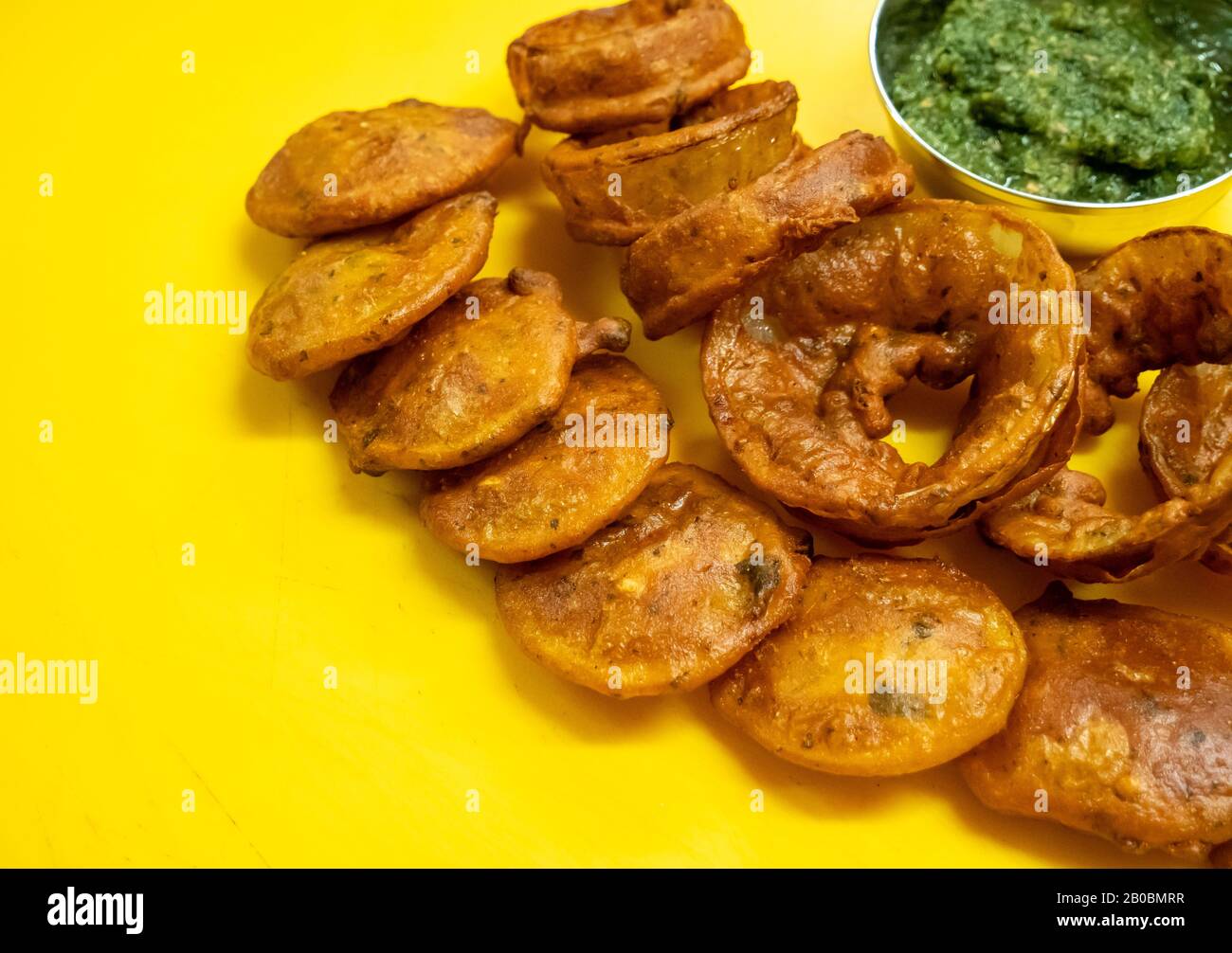 Gros plan de rondelles d'oignon et de kebab d'agneau frits indiens avec chutney à la menthe et aux herbes sur une planche à découper jaune vif. Banque D'Images