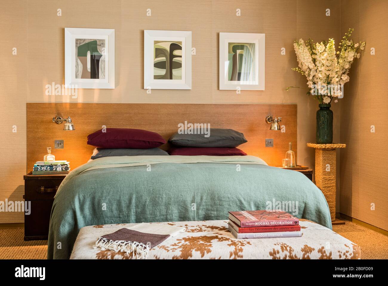L'art moderne au-dessus d'un lit dopuble avec une couverture vert clair et des livres sur un tabouret de cowhide. Banque D'Images