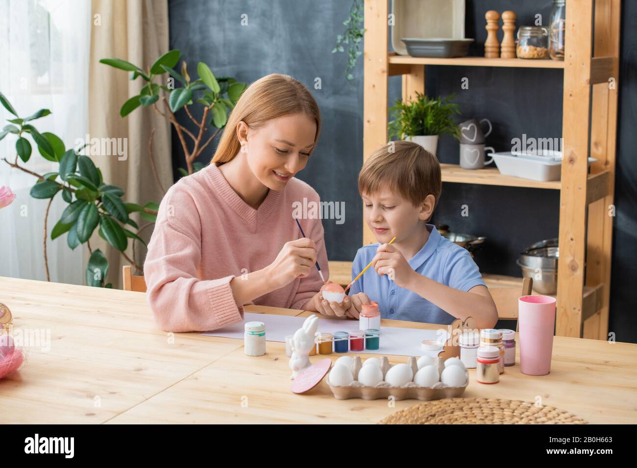 Curieux garçon assis à une table en bois et peindre des œufs avec gouache tout en passant du temps avec la mère Banque D'Images