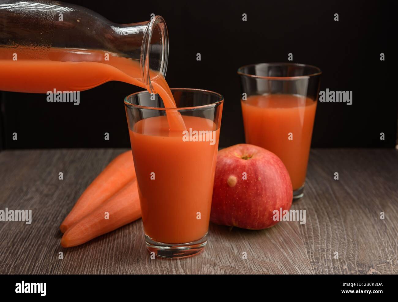 Le jus de pomme de carotte est versé de la bouteille dans un verre. Il y a des carottes et des pommes sur la table. Banque D'Images