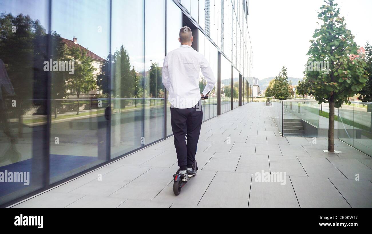 Jeune homme d'affaires décontracté qui monte en scooter électrique sur un trottoir dans une ville d'affaires Banque D'Images