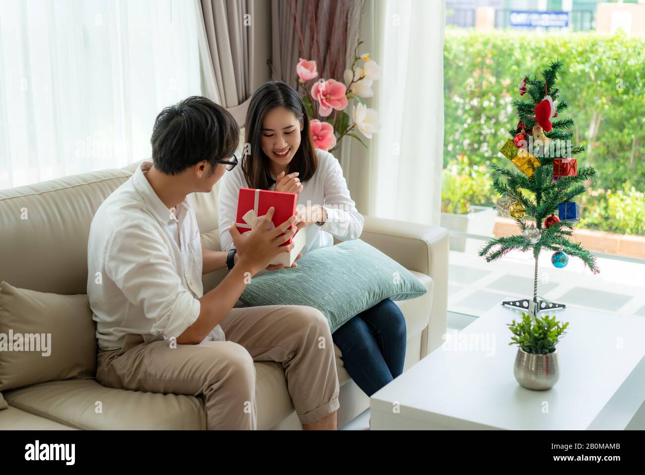 L'homme asiatique donne la femme une boîte cadeau rouge dans lequel il y a  un flacon de parfum. La femme a été ramassé sur le canapé à la maison.  L'arbre de Noël