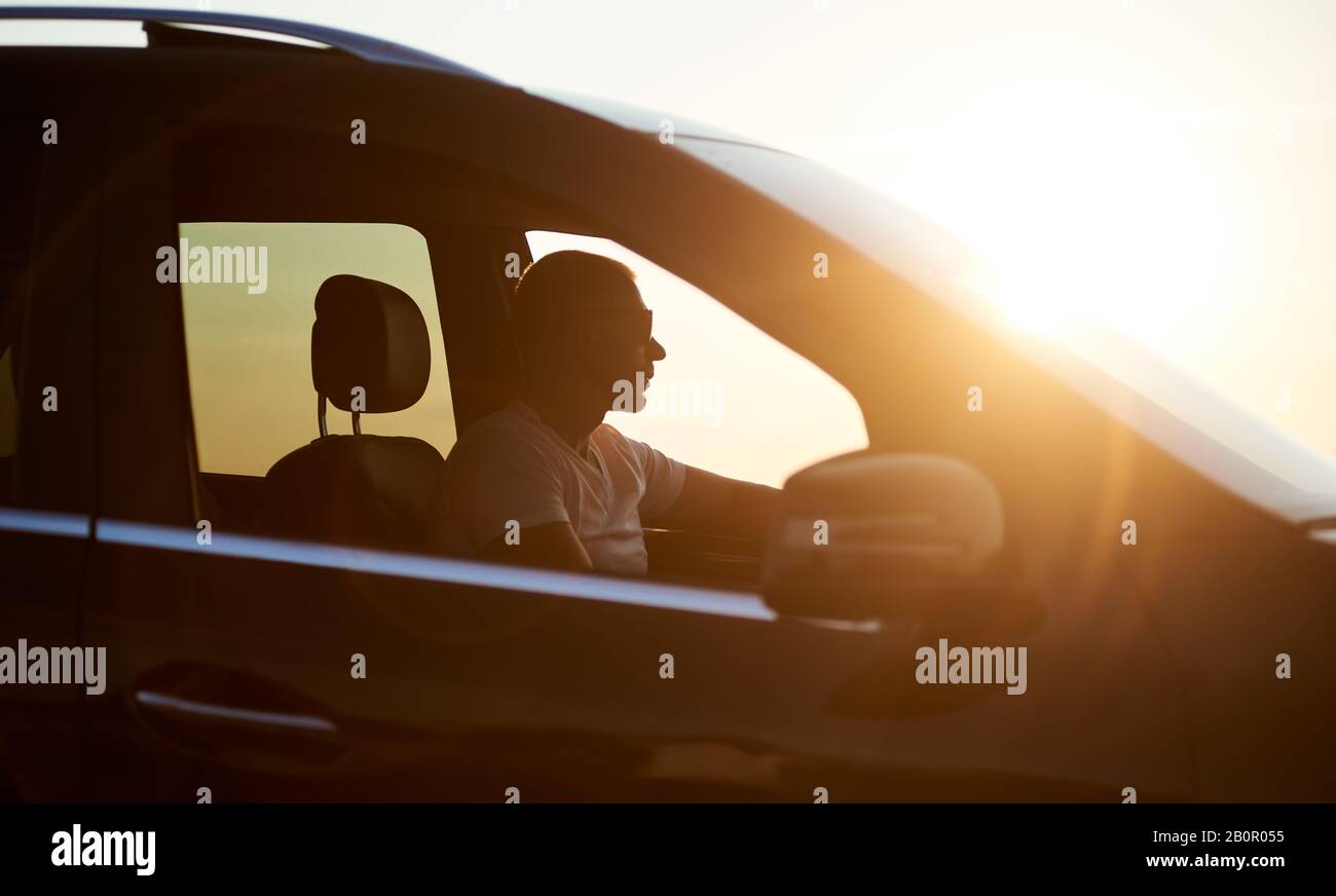 Silhouette d'un jeune homme sérieux assis dans sa voiture, portant des lunettes de soleil et regardant le coucher du soleil, soleil brillant sur la voiture, gros plan, vue latérale Banque D'Images