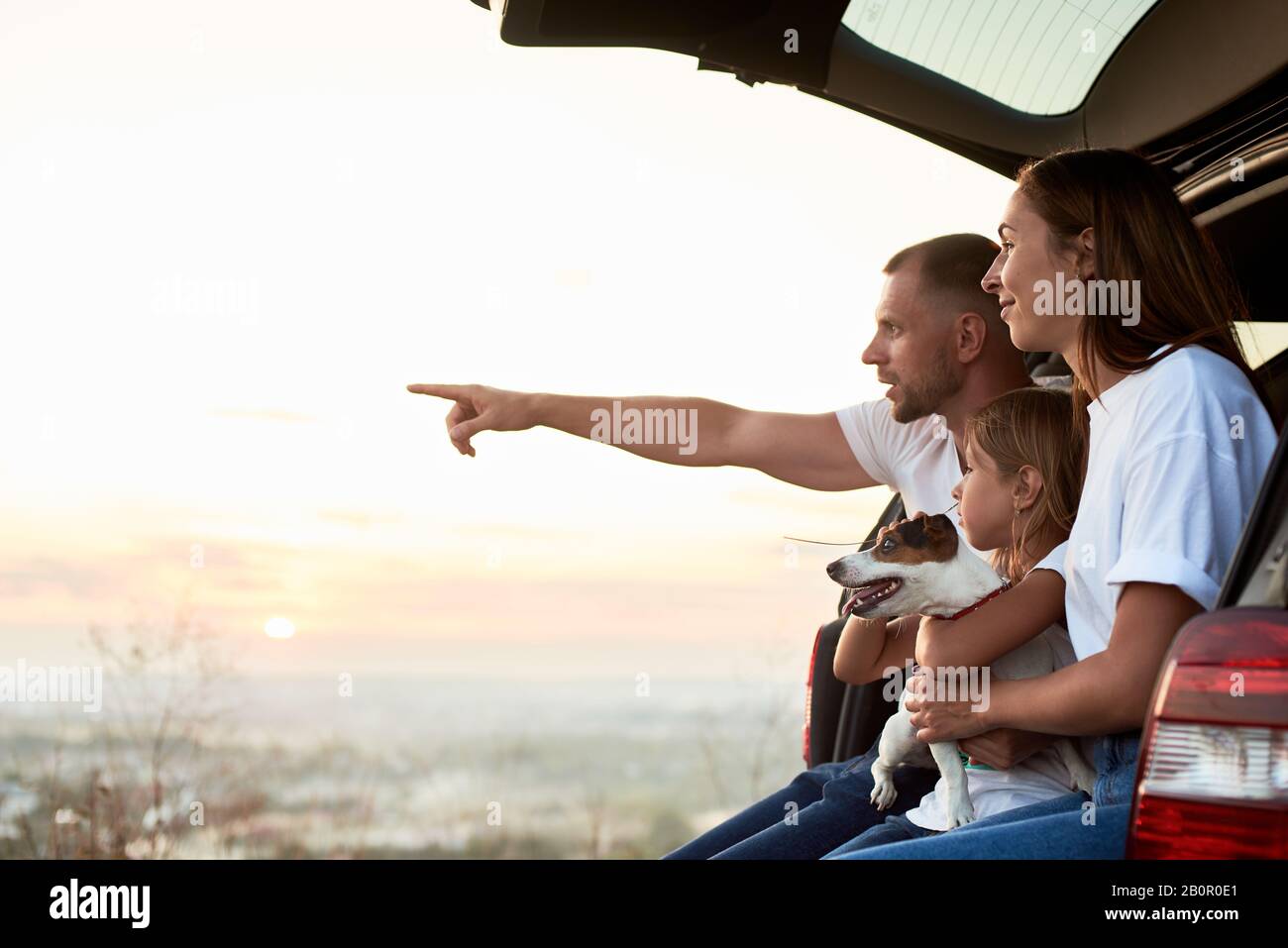 Vue latérale de la famille assise dans le coffre de voiture à l'extérieur de la ville, en regardant le coucher du soleil, le père pointe à l'horizon, copier l'espace Banque D'Images