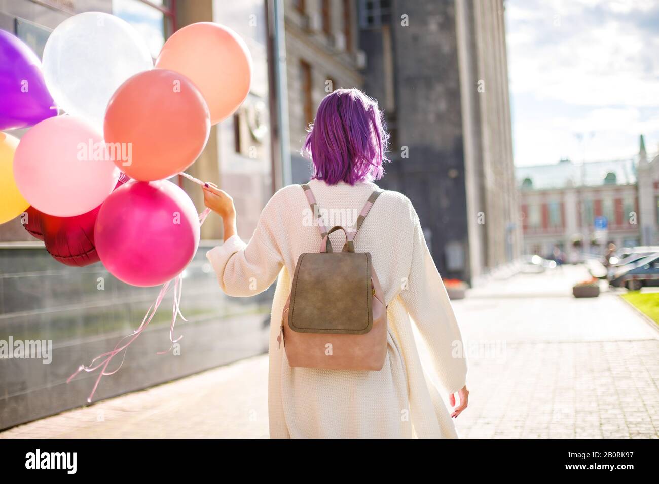 Fille aux cheveux violets, dans un gilet blanc et un sac à dos beige, avec un tas de ballons de couleur, debout la tourner en arrière devant le fond gris mur o Banque D'Images