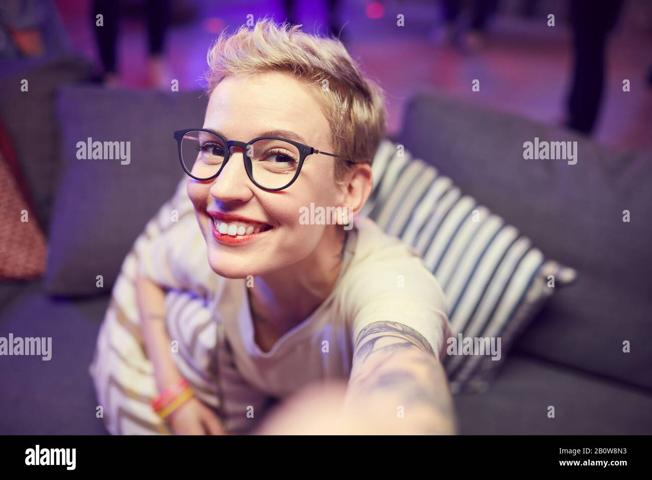 Portrait de la jeune femme avec des cheveux blonds et dans des lunettes souriant à l'appareil photo tout en étant assis sur un canapé Banque D'Images