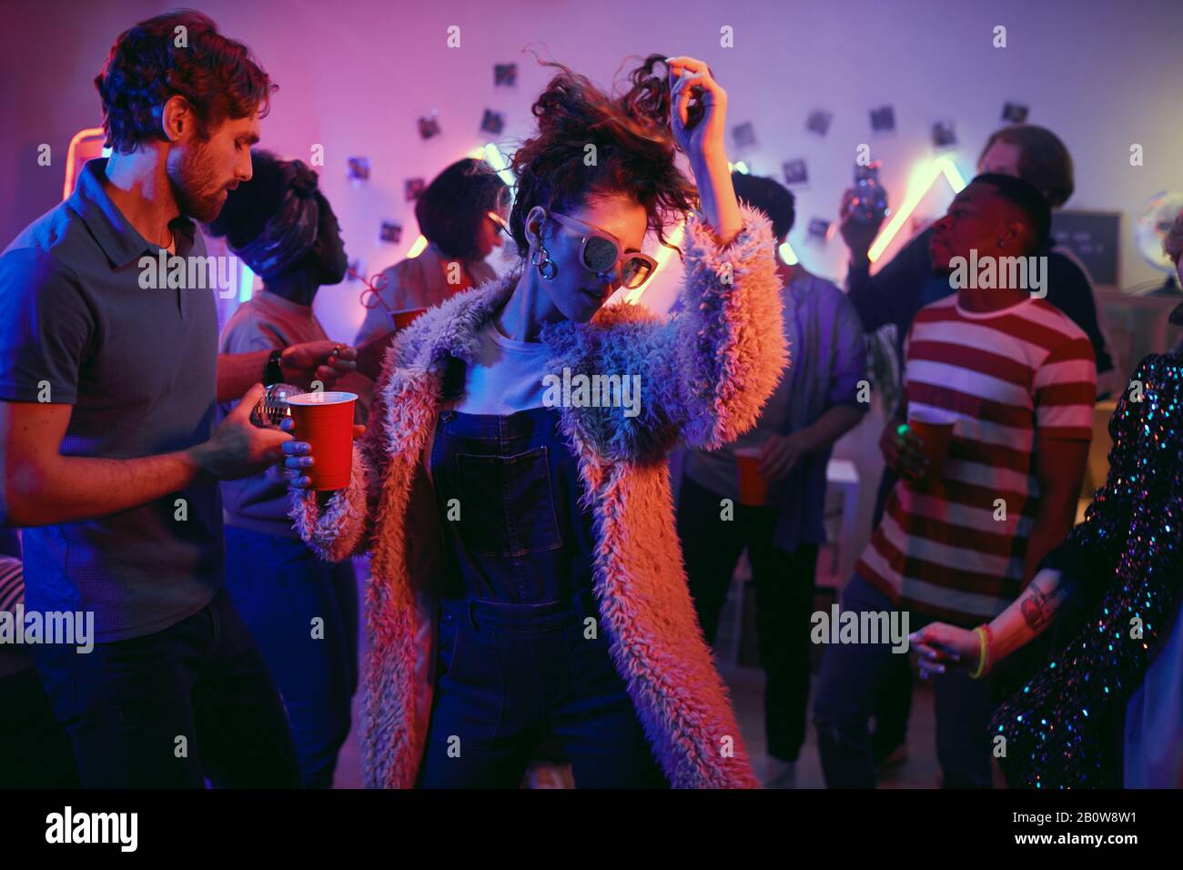 Jeune femme de mode avec boisson dansant parmi les jeunes à la fête Banque D'Images