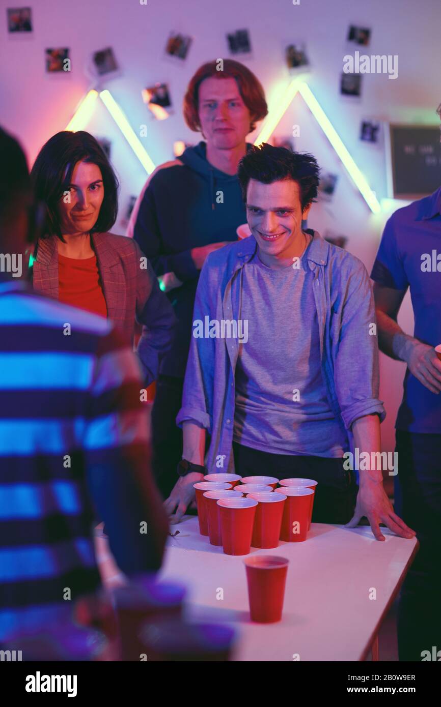 Jeune homme souriant debout près de la table avec des verres et regardant la partie de bière-pong pendant la fête Banque D'Images