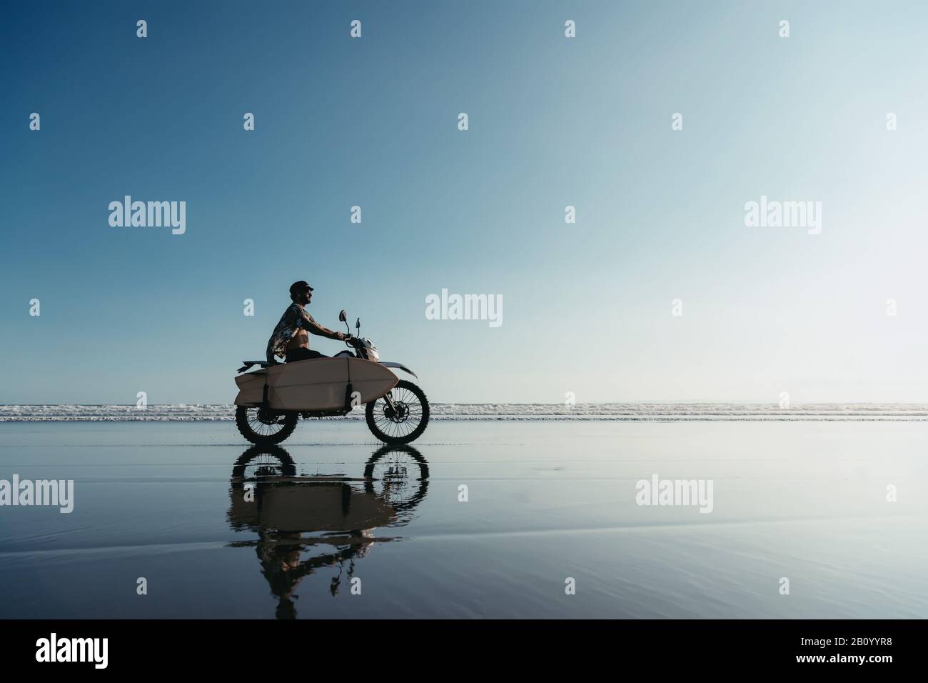 Vrai surfeur avec des promenades en planche à bord en moto enduro sur la plage de l'océan. Île de Bali, Indonésie Banque D'Images