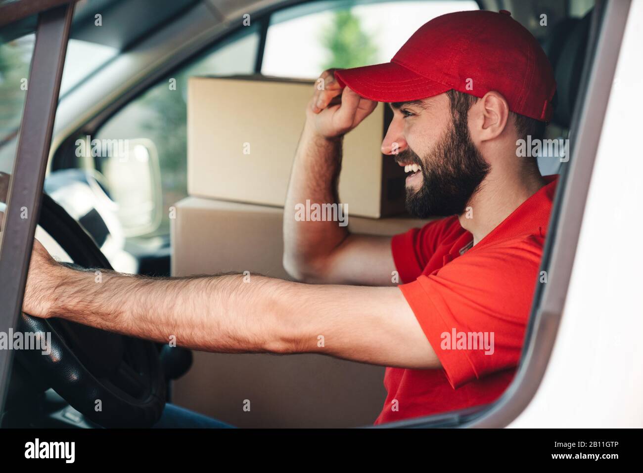 Image de l'homme de livraison barbu heureux en uniforme rouge souriant et camionnette avec des boîtes de colis Banque D'Images