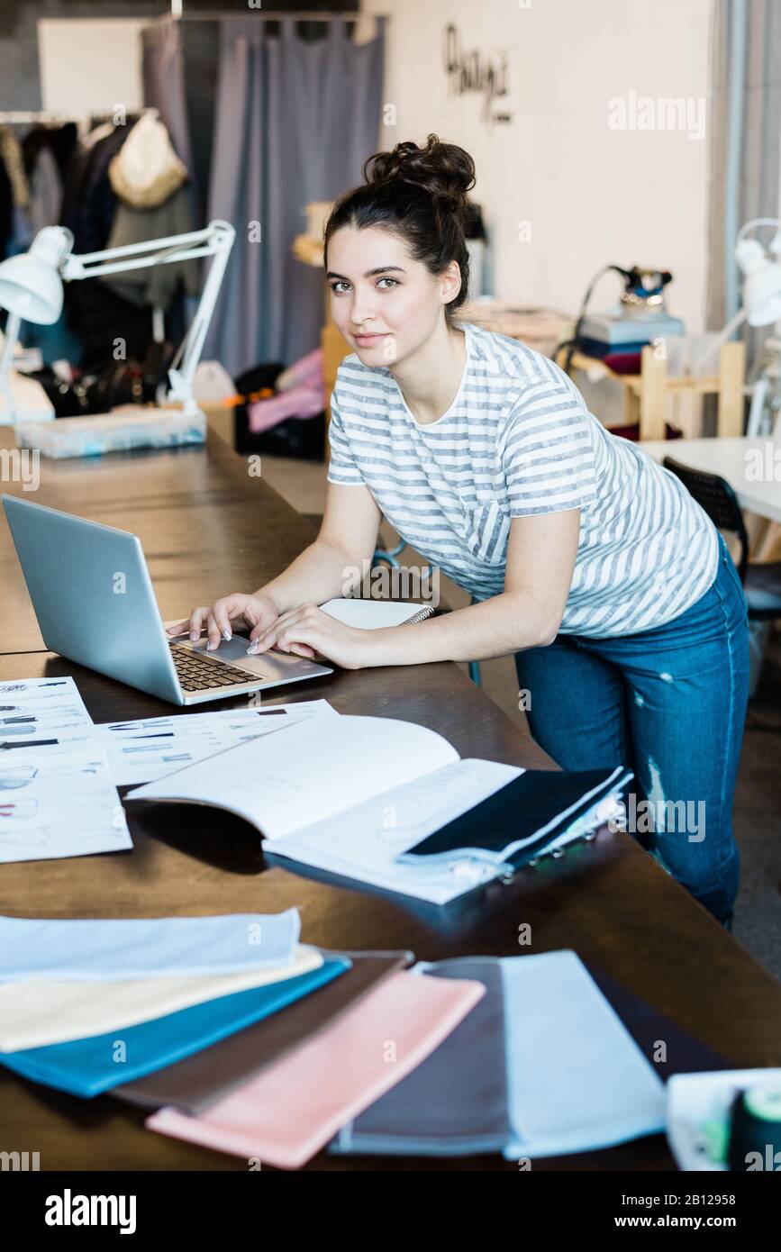 Jeune dessinateur de mode brunette en vêtements décontractés se courbant sur la table et l'ordinateur portable Banque D'Images