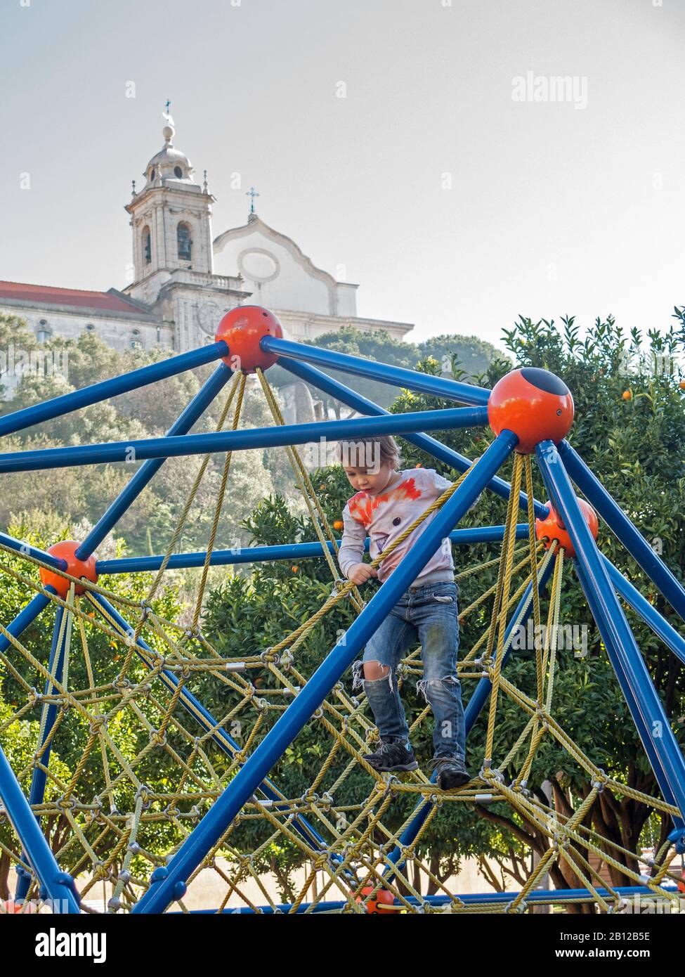 Un garçon monte sur des cordes d'un rack entre les arbres du mandarin dans le parc Jardim da Cerca. En arrière-plan, il y a une tour d'horloge de Convento de Nossa Senhora. Banque D'Images