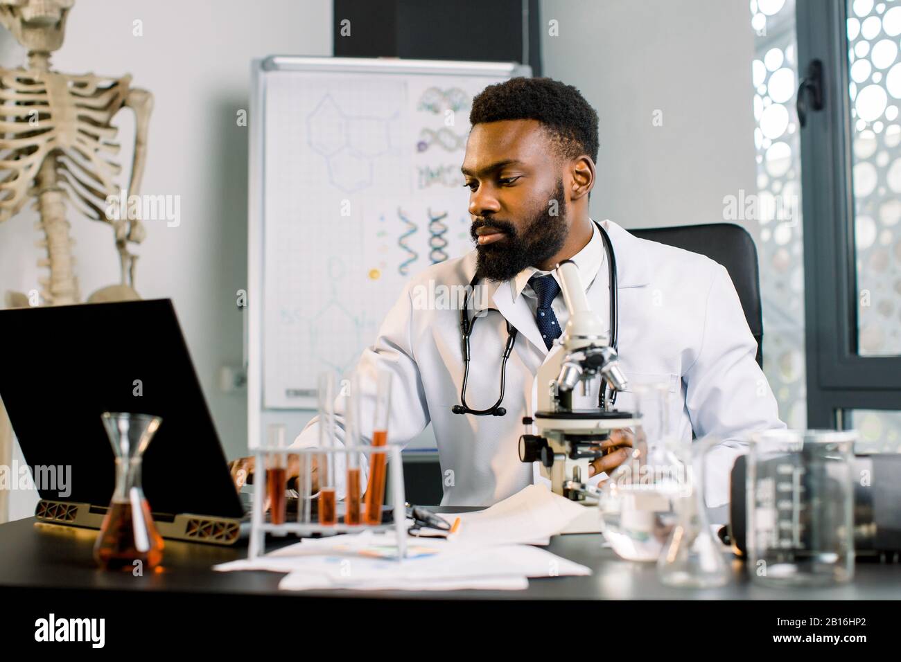 Médecin scientifique afro-américain en blouse blanche travaillant au microscope, tubes à essai en laboratoire, prenant des notes sur l'ordinateur portable. Biotechnologie, médecine, im Banque D'Images