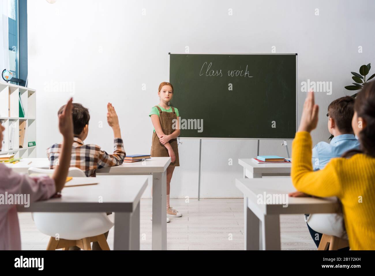 foyer sélectif de l'écolière debout près du tableau de surveillance avec lettrage de travail de classe et des camarades de classe avec des mains levées Banque D'Images