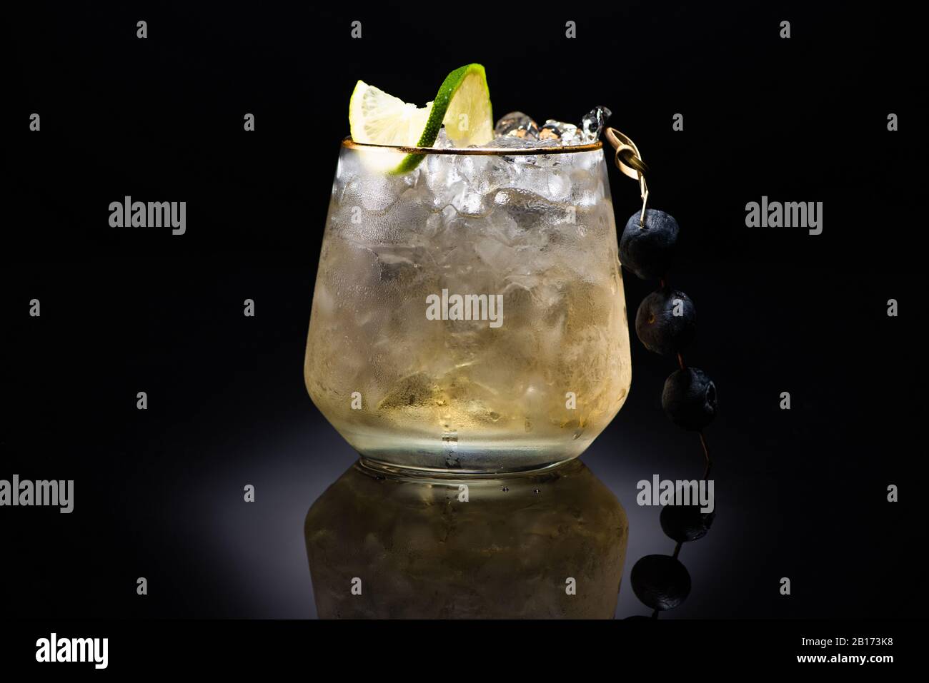 verre transparent avec glace et liquide doré garni de chaux et de bleuets sur fond noir Banque D'Images