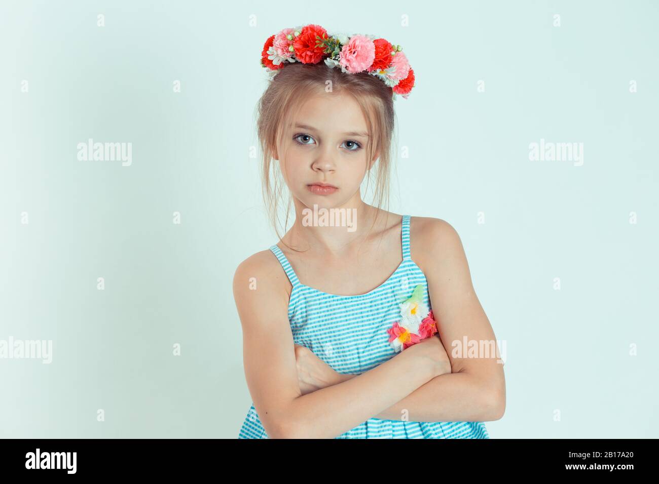 Jeune fille canneuse regardant la caméra avec les bras croisés, les mains repliées. Gros plan portrait de modèle caucasien pour enfant avec bandeau floral isolé sur blanc vert gr Banque D'Images
