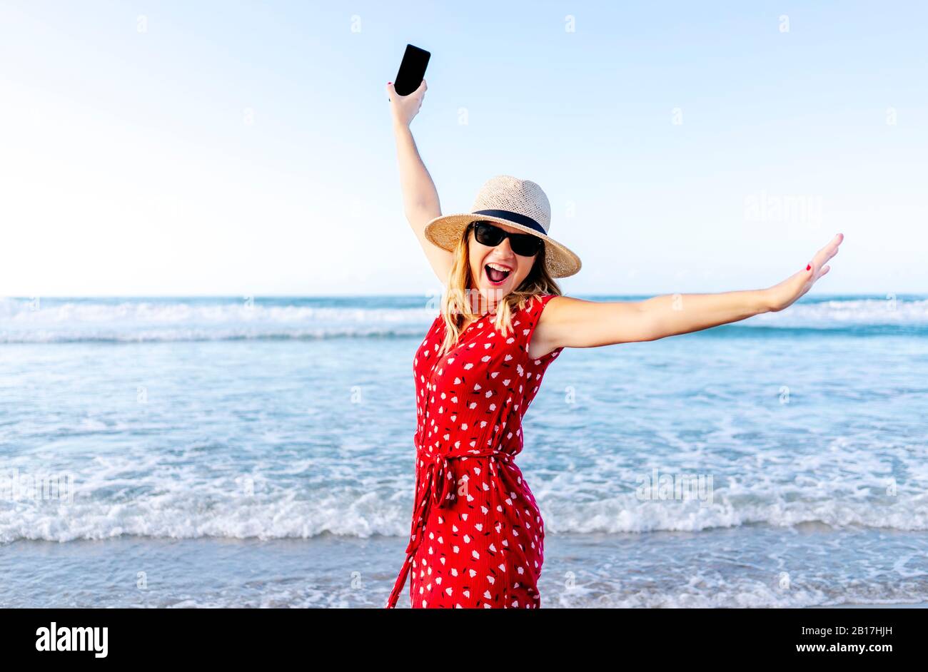 Femme blonde souriante portant une robe rouge et un chapeau et tenant son smartphone à la plage Banque D'Images