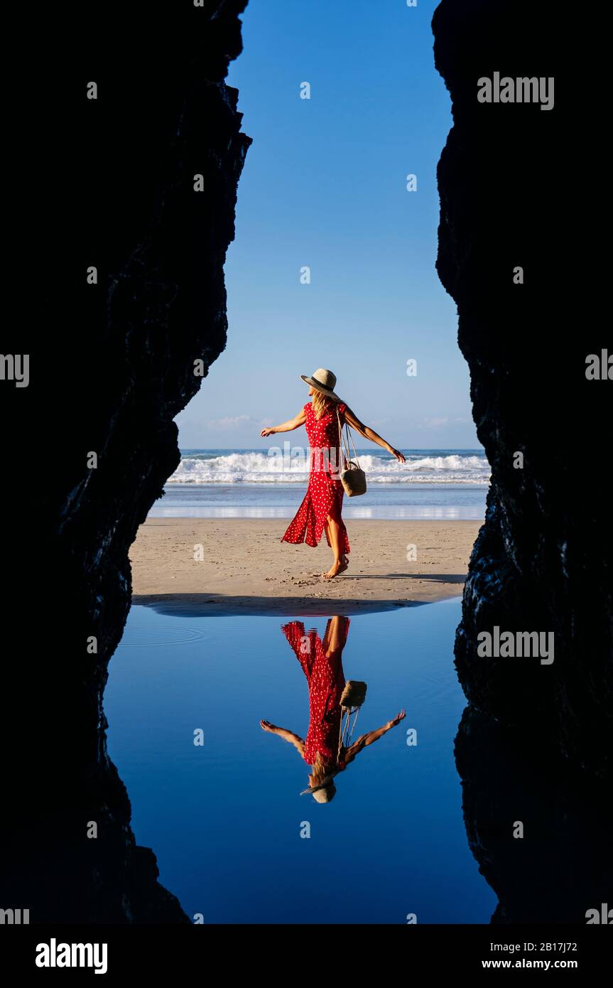 Blonde femme portant robe rouge et chapeau et danse à la plage, Playa de Las Catedrales, Espagne Banque D'Images