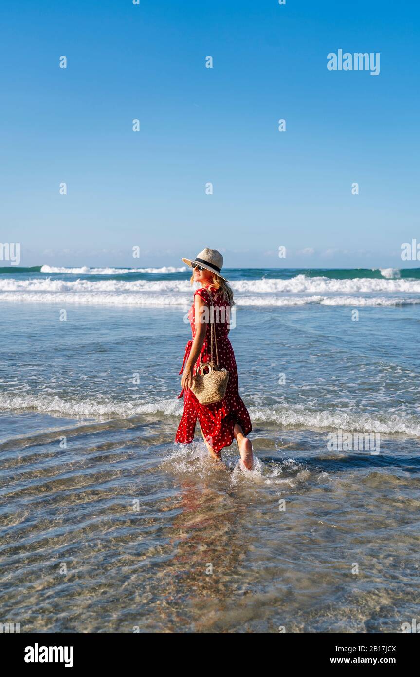 Blonde femme portant une robe rouge et chapeau et marchant à la plage, Playa de Las Catedrales, Espagne Banque D'Images