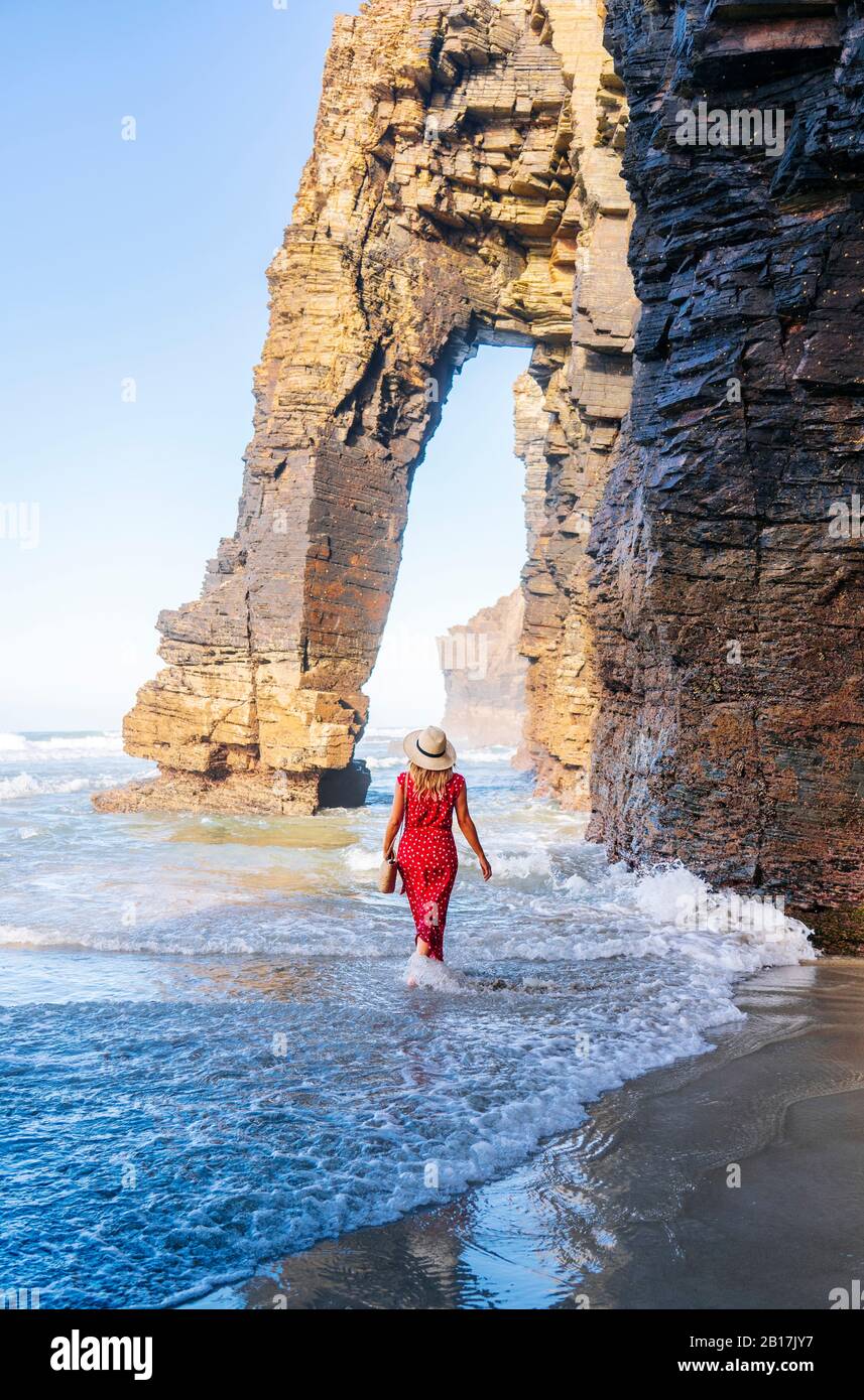 Blonde femme portant robe rouge et chapeau et marchant le long de la plage, Arche naturelle à Playa de Las Catedrales, Espagne Banque D'Images