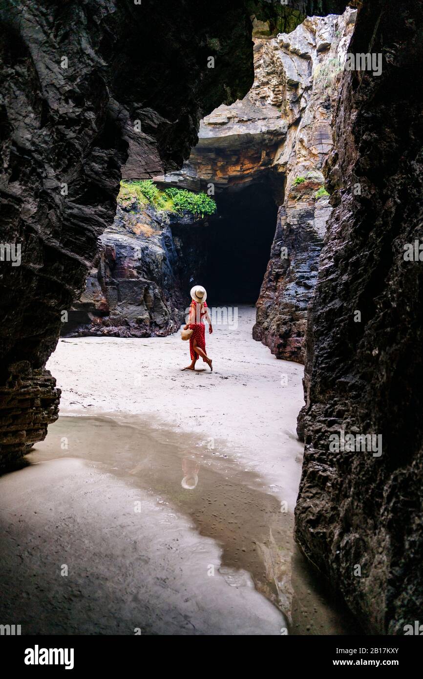 Blonde femme portant une robe rouge et chapeau dans une grotte de roche, Playa de Las Catedrales, Espagne Banque D'Images