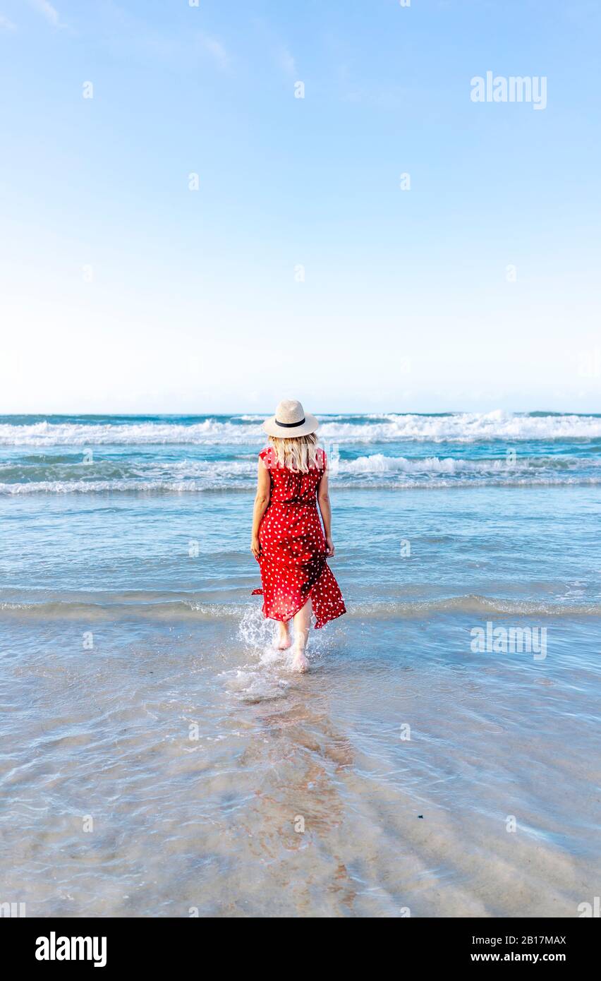 Vue arrière d'une femme blonde portant une robe rouge et un chapeau et marchant sur la plage dans l'eau Banque D'Images