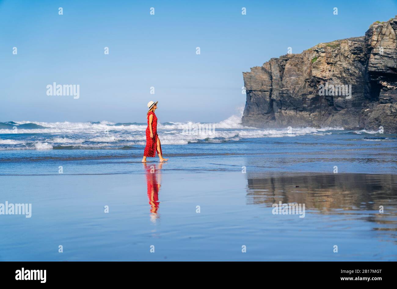 Blonde femme portant une robe rouge et chapeau et marchant le long de la plage, Playa de Las Catedrales, Espagne Banque D'Images