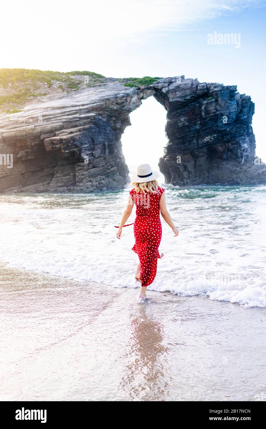 Blonde femme portant une robe rouge et chapeau et marchant à la plage, Natural Arch à Playa de Las Catedrales, Espagne Banque D'Images