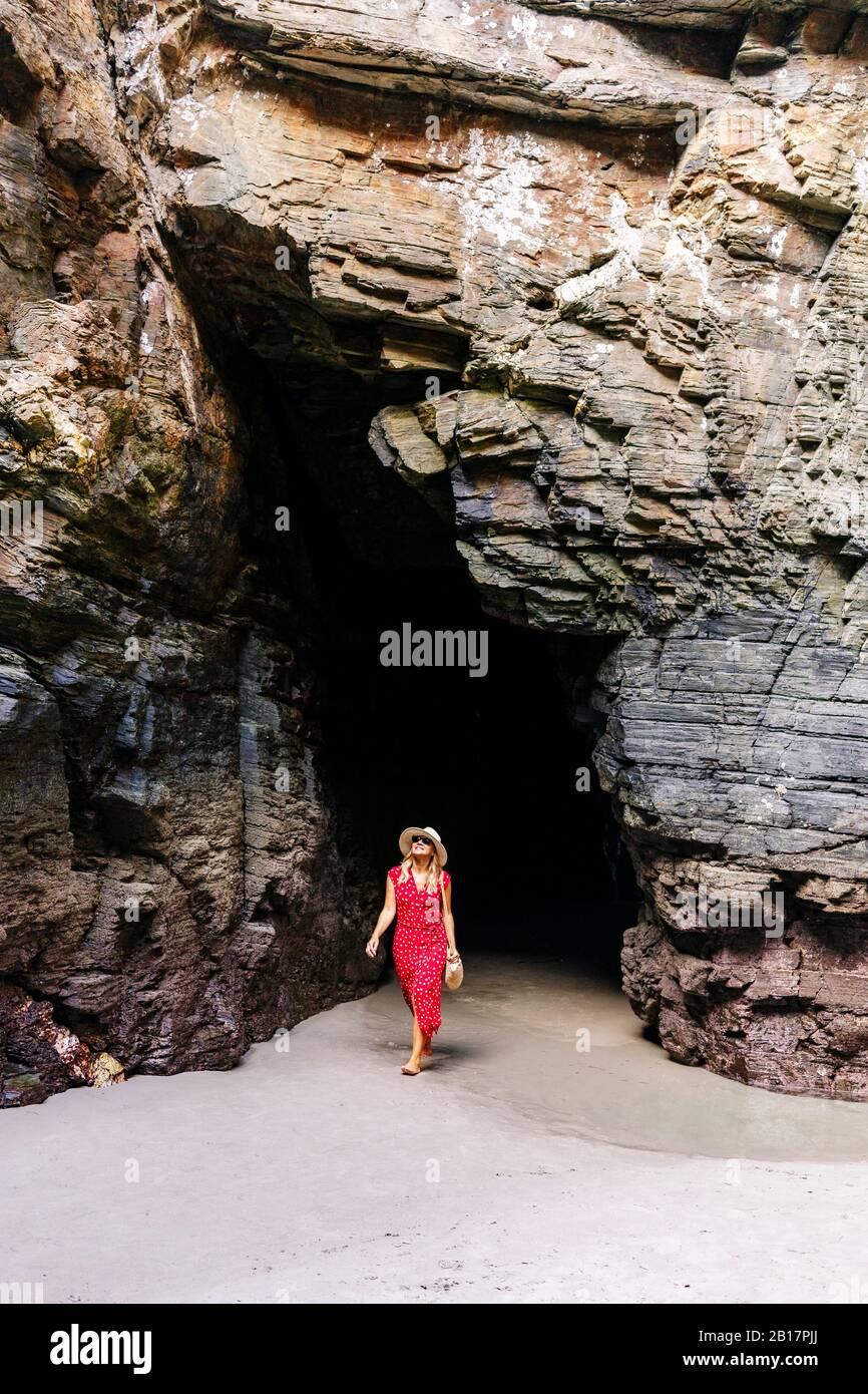 Blonde femme portant une robe rouge et chapeau dans une grotte de roche, Playa de Las Catedrales, Espagne Banque D'Images