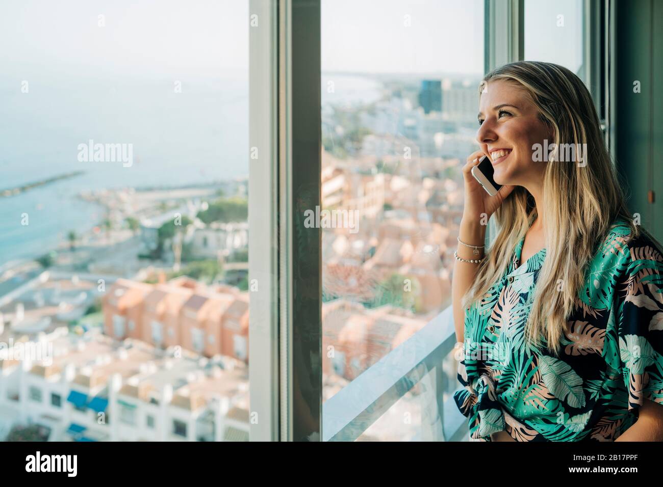 Femme souriante au téléphone à la fenêtre de la ville côtière Banque D'Images
