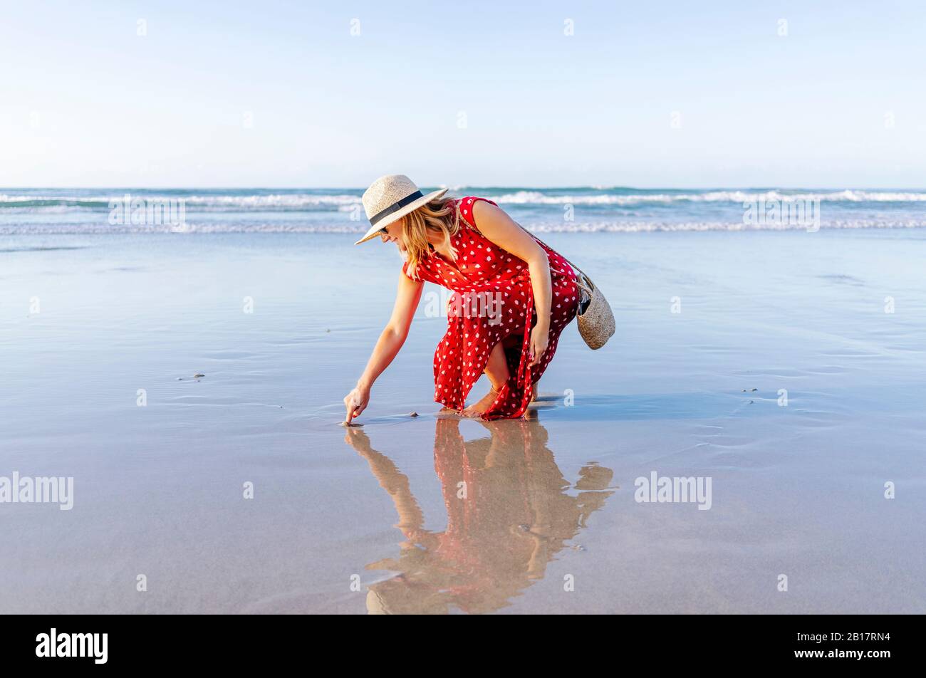 Blonde femme portant une robe rouge et chapeau à la plage, Playa de Las Catedrales, Espagne Banque D'Images