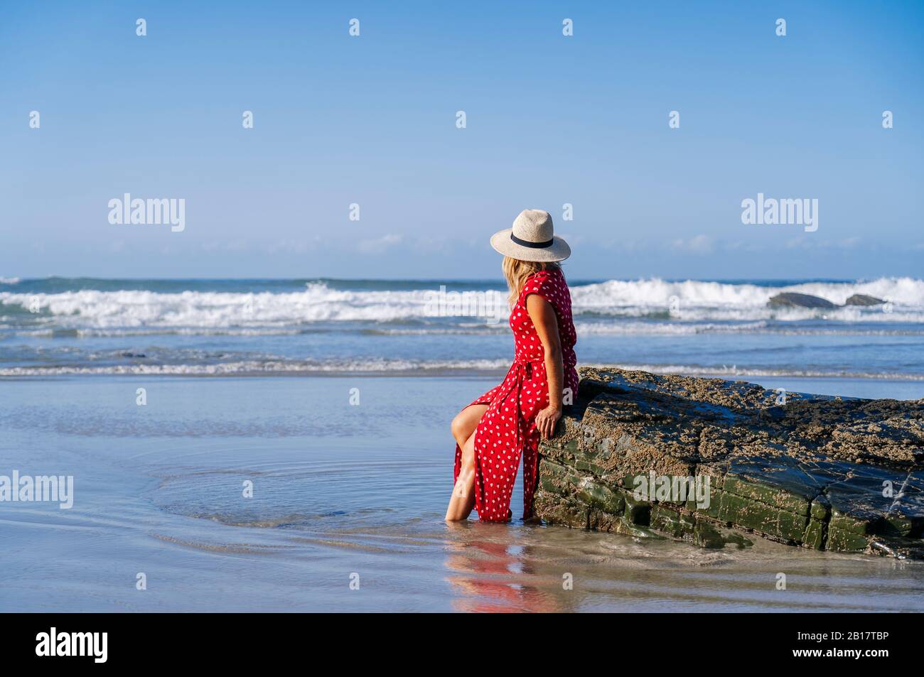 Blonde femme portant robe rouge et chapeau sittig sur le rocher à la plage, Playa de Las Catedrales, Espagne Banque D'Images