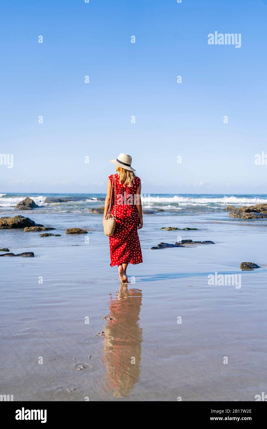 Blonde femme portant une robe rouge et chapeau et marchant le long de la plage, Playa de Las Catedrales, Espagne Banque D'Images