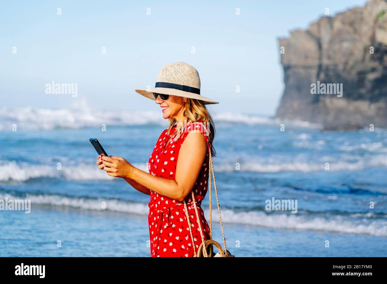 Blonde femme portant une robe et un chapeau rouges et utilisant un smartphone à la plage Banque D'Images