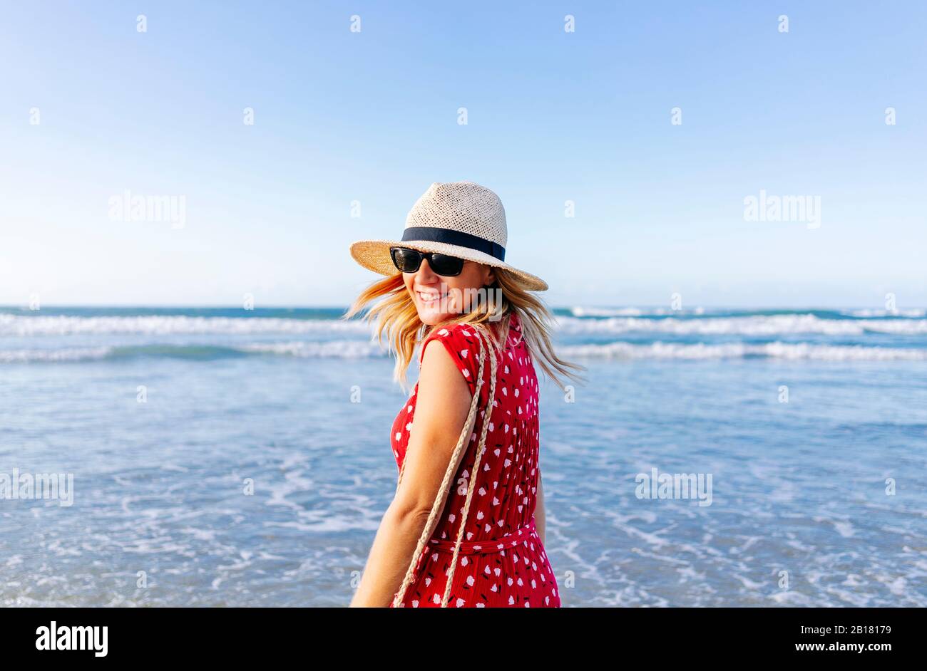 Blonde femme portant une robe rouge et chapeau à la plage, tournant et regardant l'appareil photo Banque D'Images