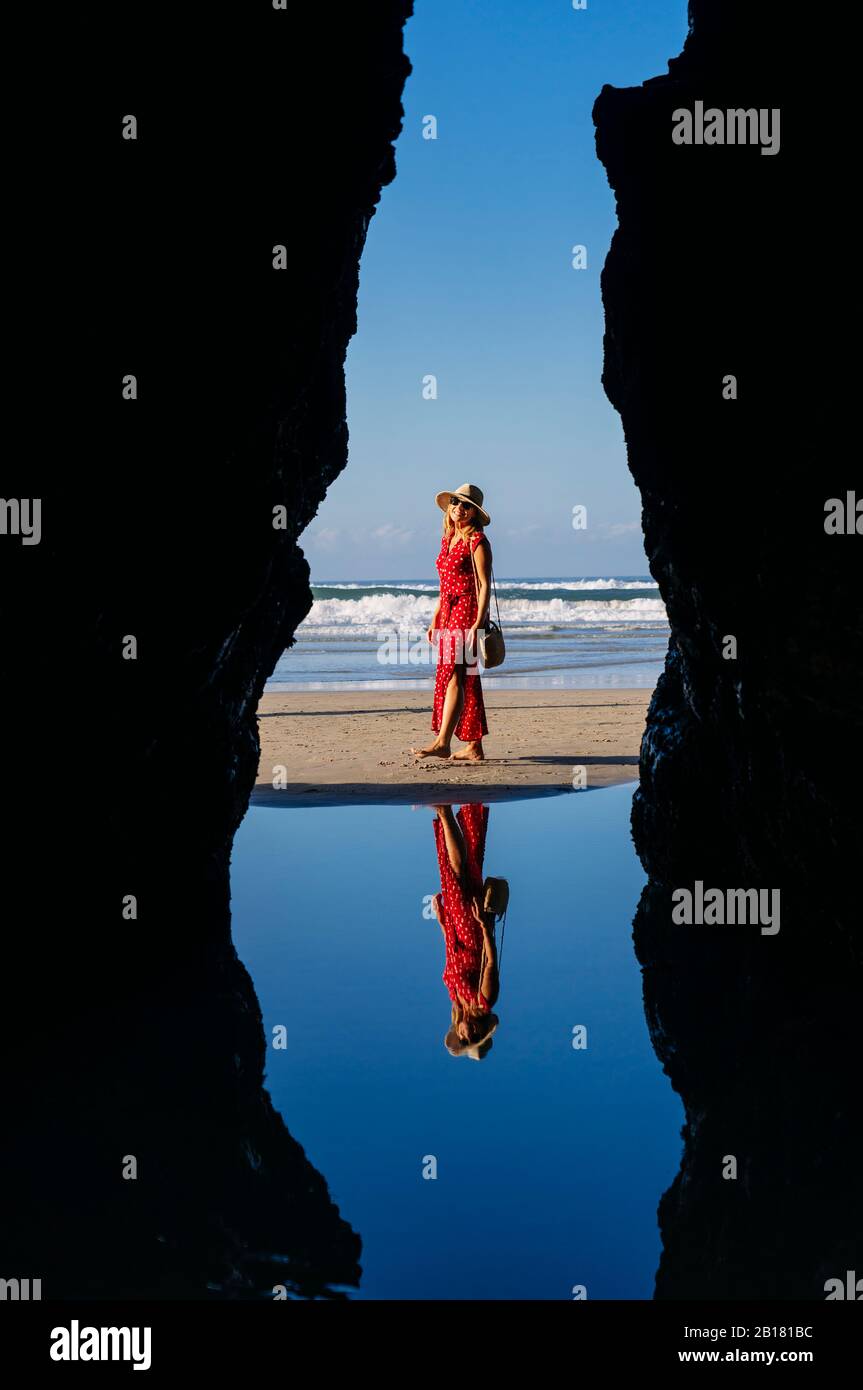 Blonde femme portant robe rouge et chapeau et marchant le long de la plage, Arche naturelle à Playa de Las Catedrales, Espagne Banque D'Images