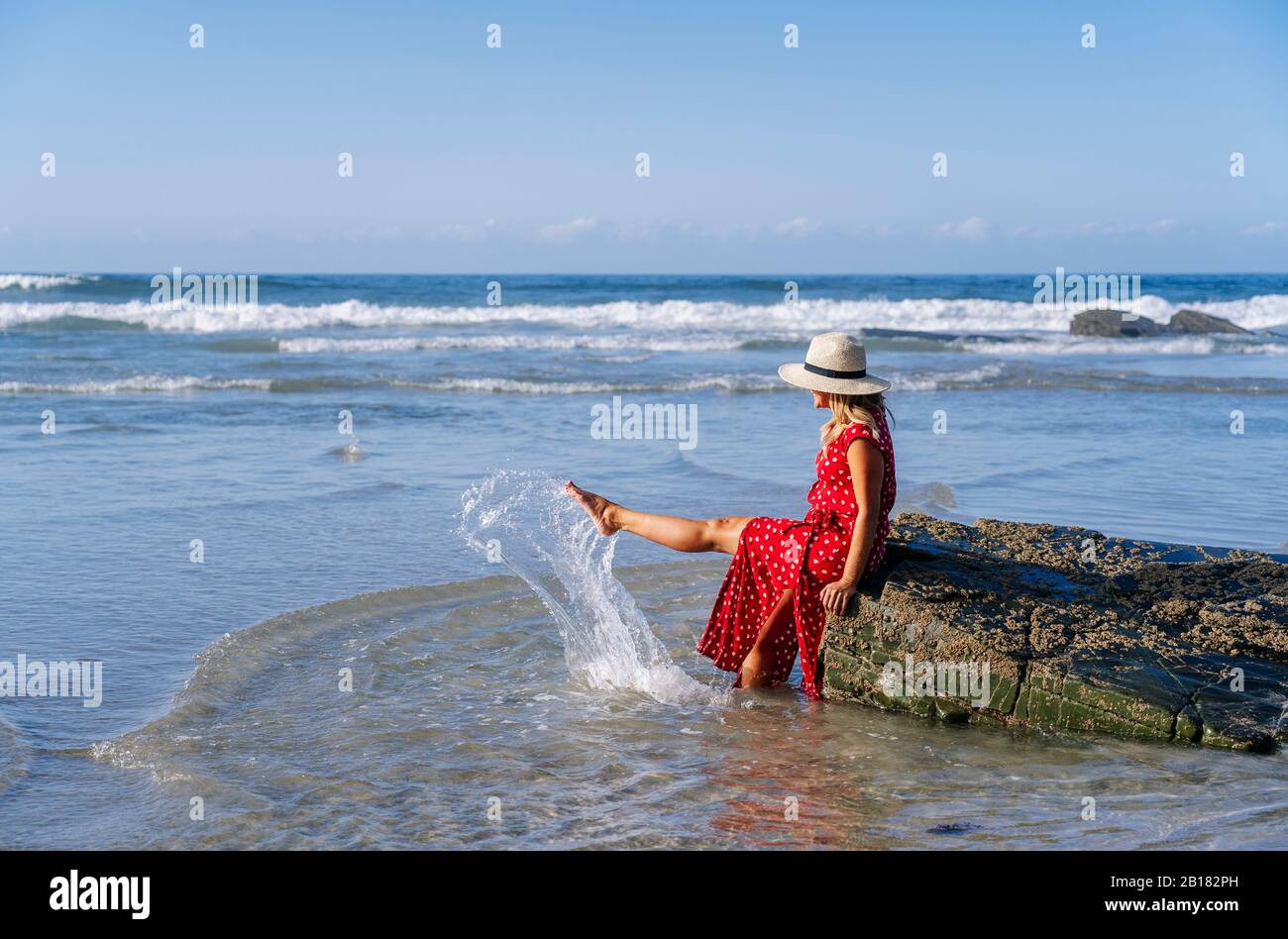 Blonde femme portant robe rouge et chapeau sittig sur le rocher à la plage, Playa de Las Catedrales, Espagne Banque D'Images