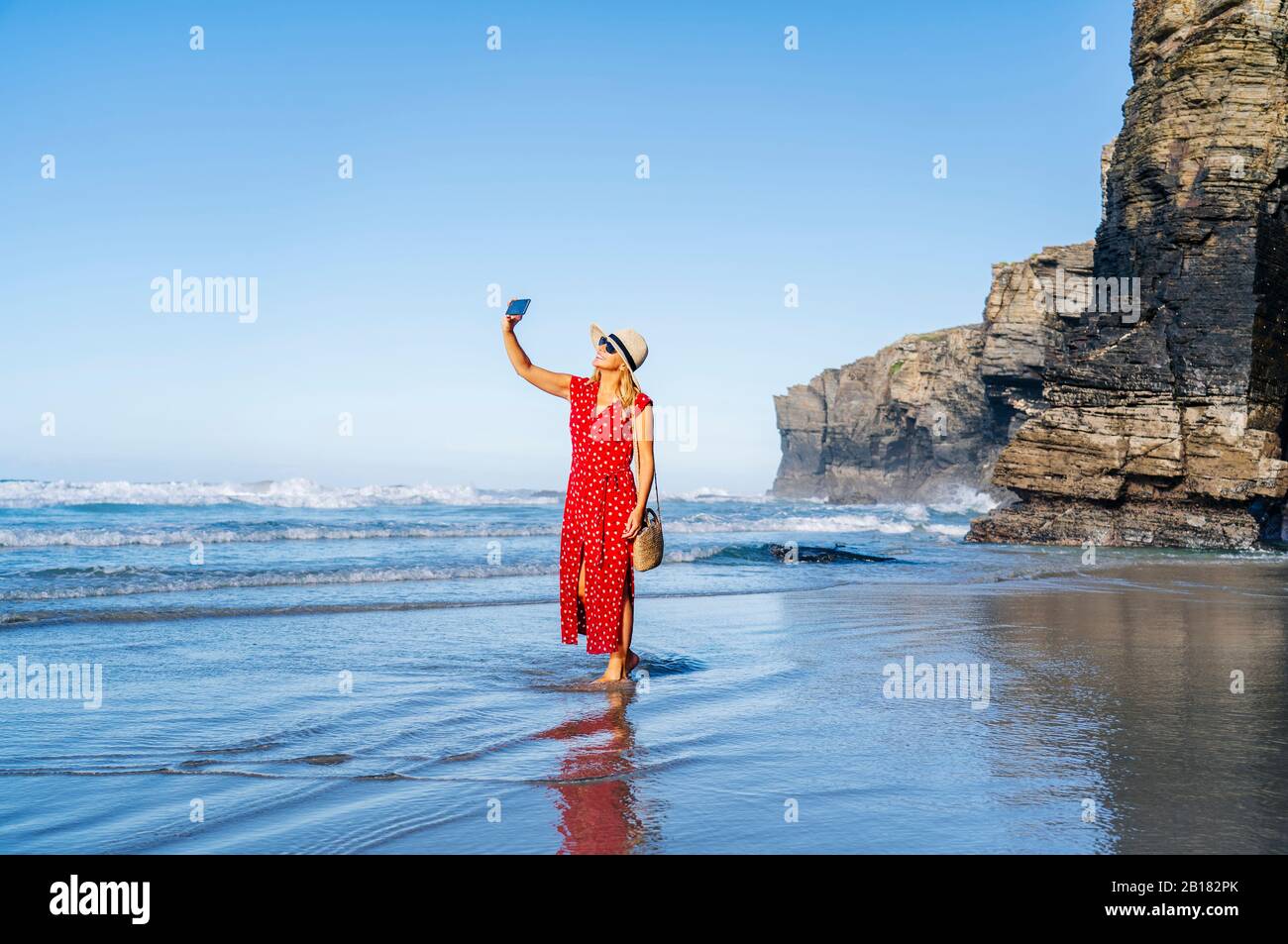 Blonde femme portant une robe et un chapeau rouges et utilisant un smartphone à la plage Banque D'Images