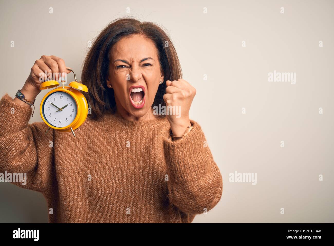 Une femme brunette d'âge moyen tenant un réveil clasique sur fond isolé en  colère et en colère, se moquant de la colère et de la furieuse, tout en  criant avec un Photo