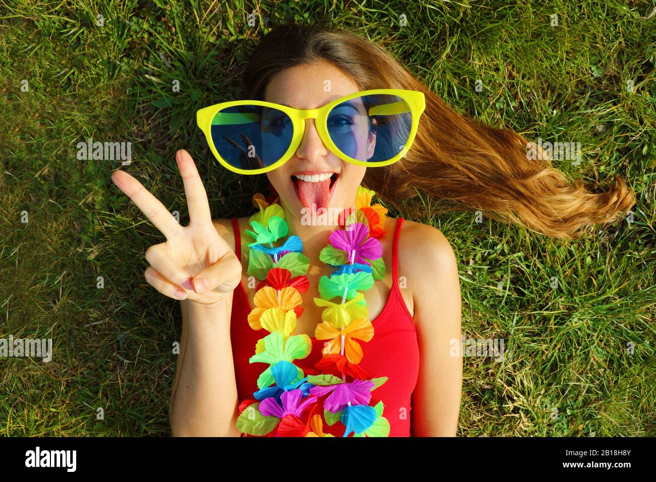 Fille après la fête du Carnaval. Jeune femme avec de grandes lunettes de  soleil drôles et une guirlande carnaval posée sur l'herbe montrant la  langue et le signe V Photo Stock -