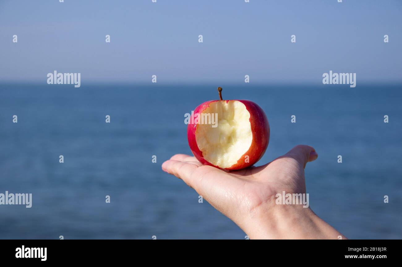 Pomme mordu rouge en main contre la mer. Écotourisme et consommation de fruits naturels et biologiques. Harmonie de concept entre loisirs et écologie. Banque D'Images