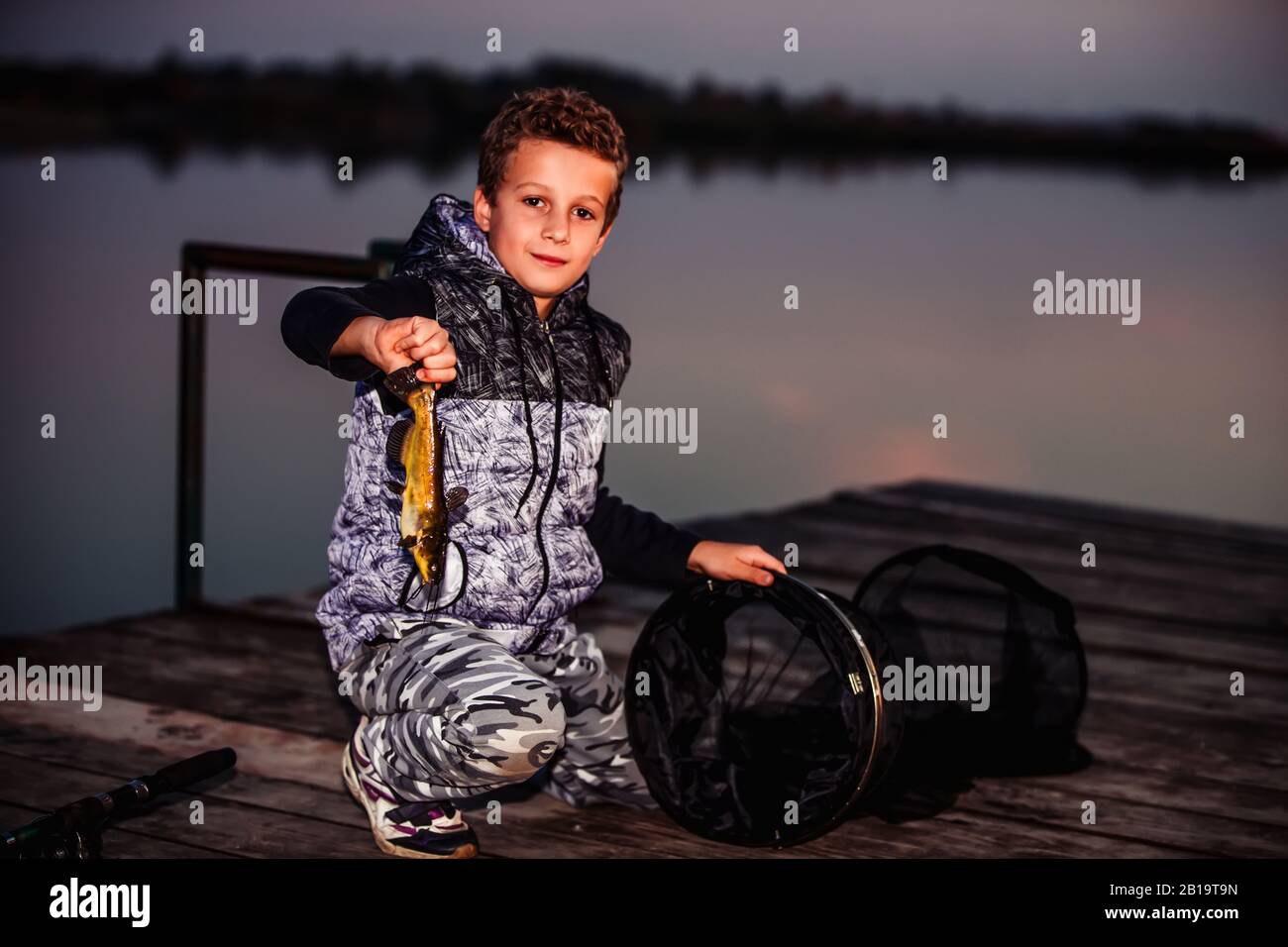 Un joli petit garçon pêchant sur un lac le soir et montrant des poissons pêchés à l'extérieur Banque D'Images