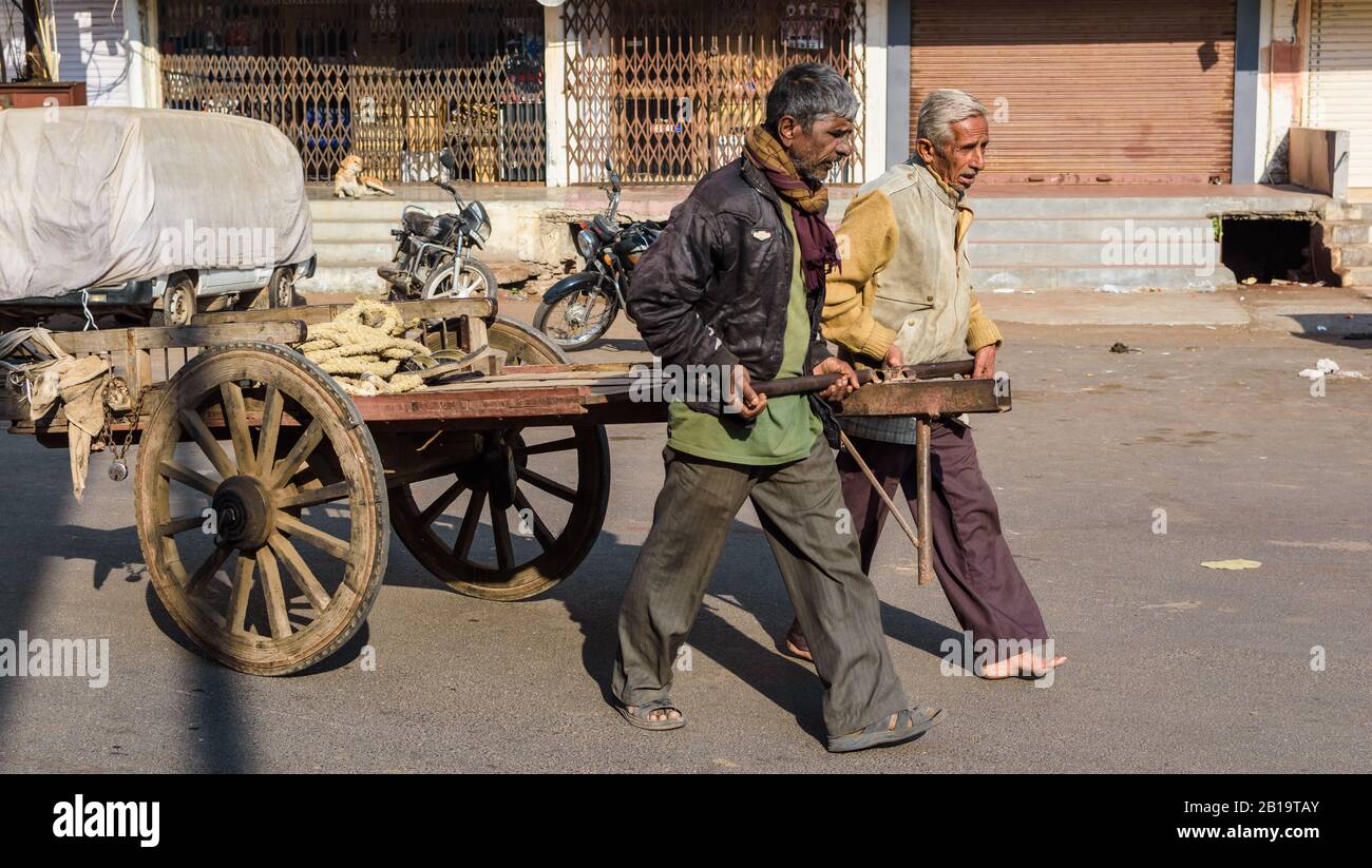 Jamnagar, Gujarat, Inde - décembre 2018 : deux ouvriers tirent leur charrette de bois sur les rues commerçantes de la ville. Banque D'Images Jamnagar, Gujarat, Inde - décembre 2018 : deux ouvriers tirent leur charrette de bois sur les rues commerçantes de la ville. Banque D'Images