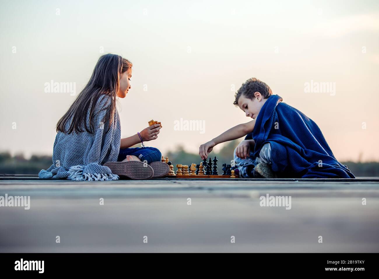 Deux jeunes petits amis mignons, un garçon et une fille qui s'amusent en jouant aux échecs assis près du lac le soir. Les enfants jouent. Amitié. Banque D'Images
