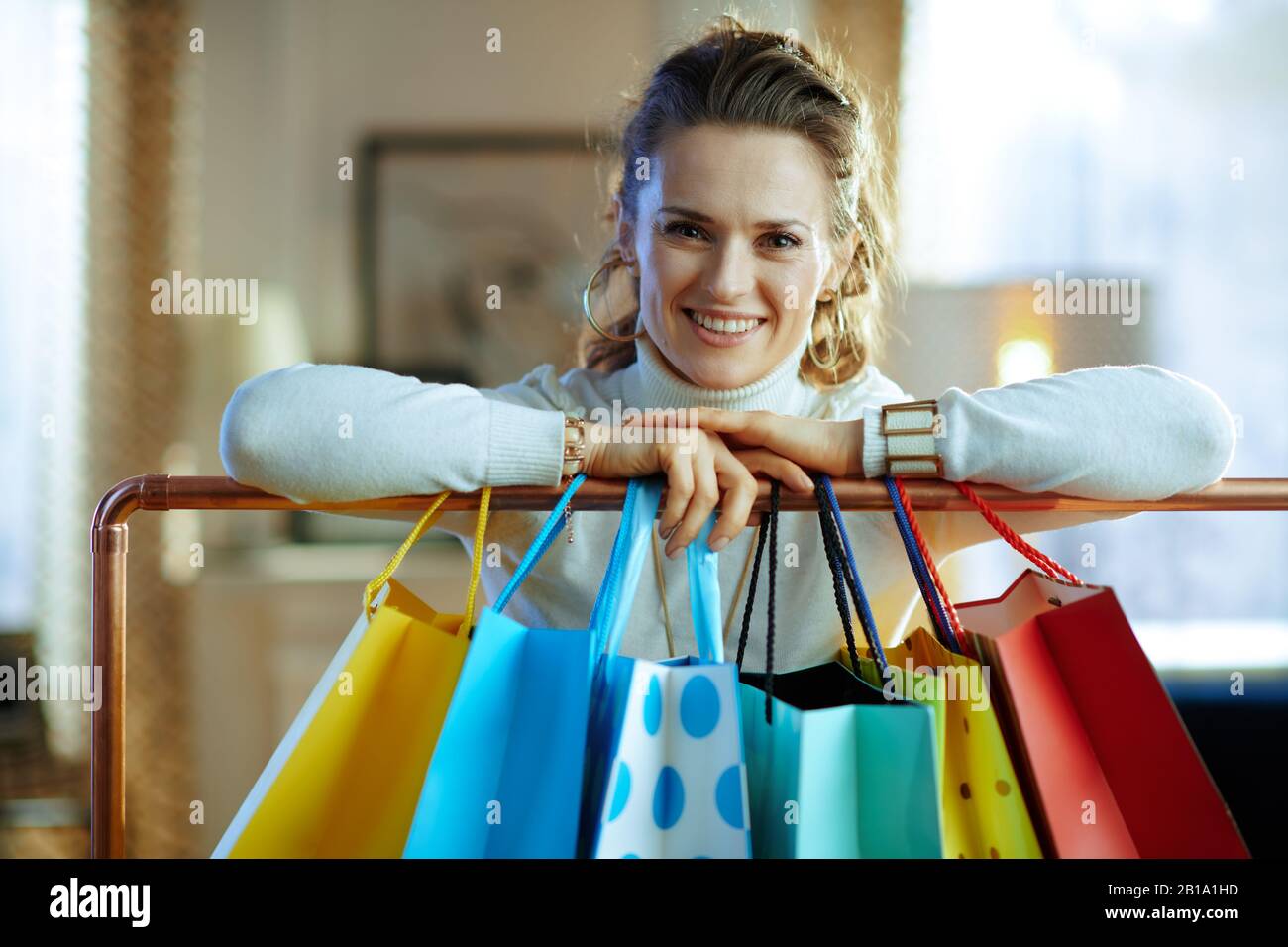 heureuse femme moderne de 40 ans en chandail blanc et jupe près des sacs de shopping colorés accrochés sur des vêtements en cuivre rail. Banque D'Images