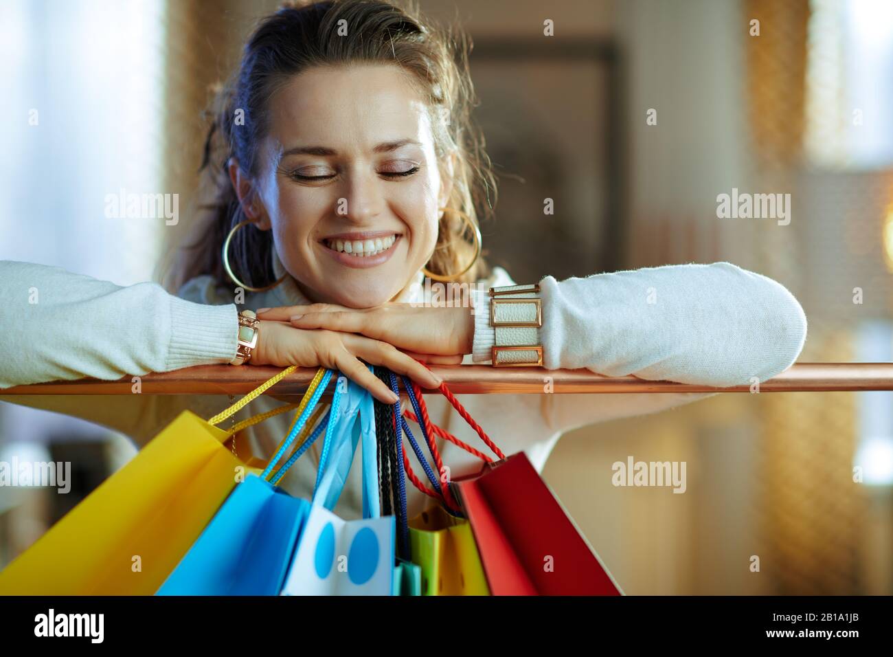 jeune femme souriante dans un pull blanc et une jupe près de sacs de shopping colorés accrochés sur un rail de vêtements en cuivre. Banque D'Images