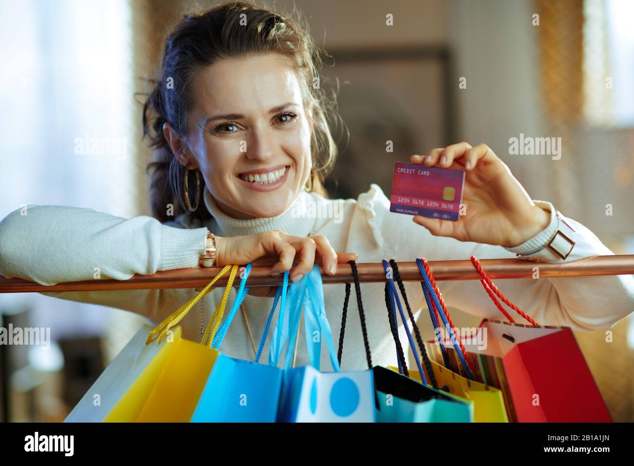 femme souriante moderne de taille moyenne dans un pull blanc et une jupe près de sacs à provisions colorés accrochés sur un rail en cuivre montrant une carte de crédit rouge. Banque D'Images