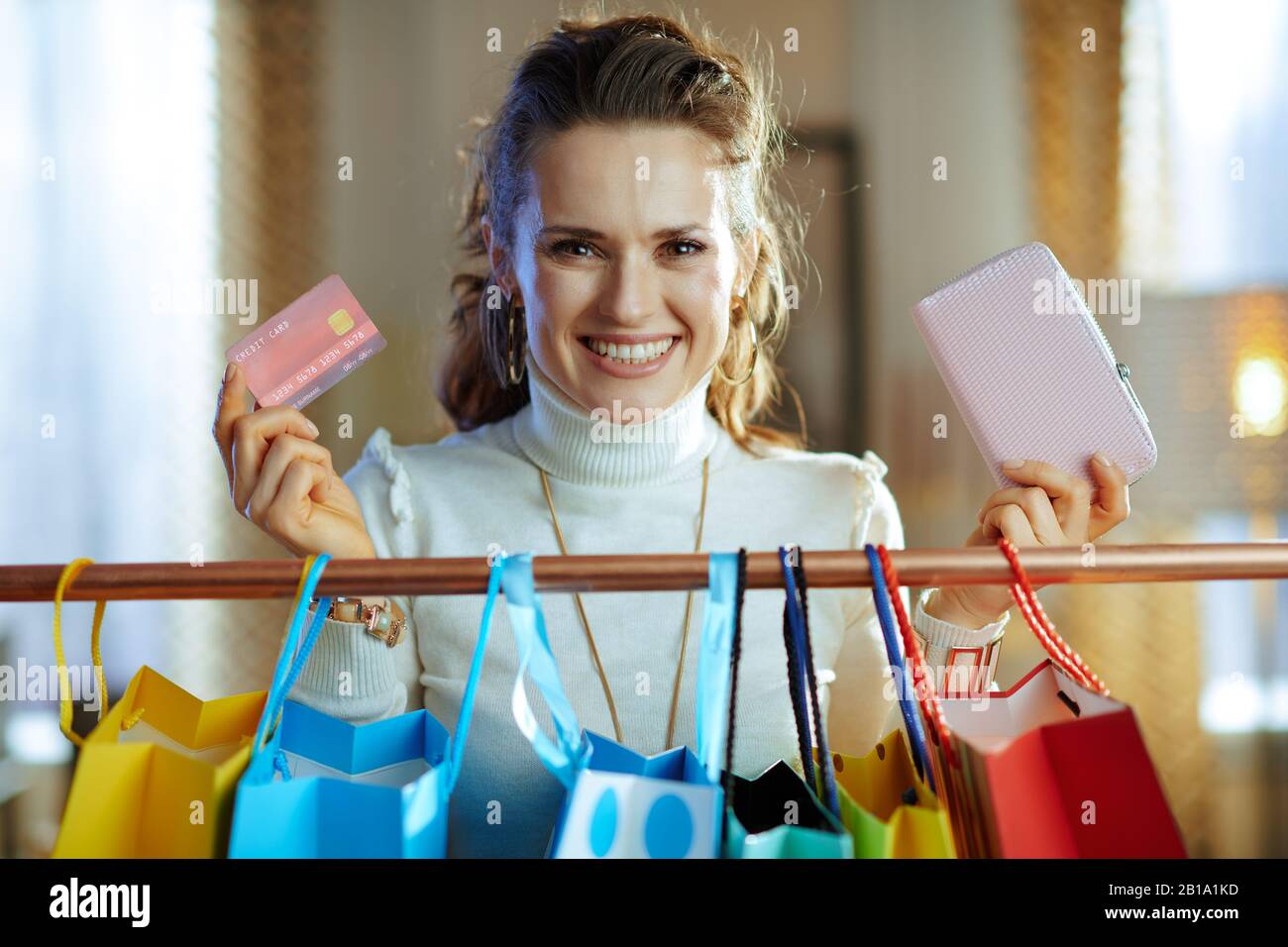 femme souriante et tendance d'âge moyen en sweat blanc et jupe avec carte de crédit et portefeuille près des sacs de shopping colorés accrochés sur des vêtements en cuivre rail. Banque D'Images