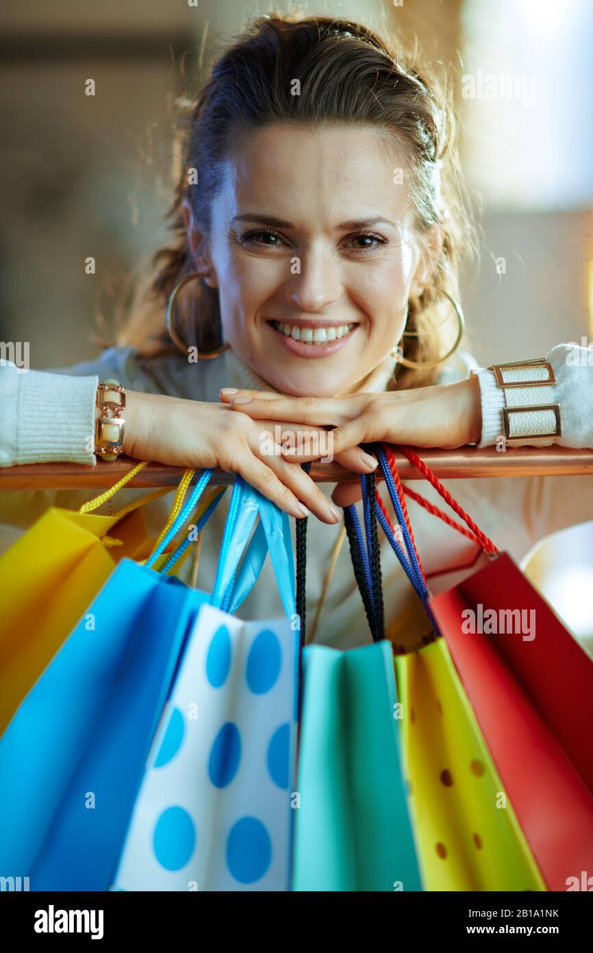 Portrait d'une heureuse femme de 40 ans à la mode dans un pull blanc et une jupe près de sacs de shopping colorés accrochés sur un rail de vêtements en cuivre. Banque D'Images