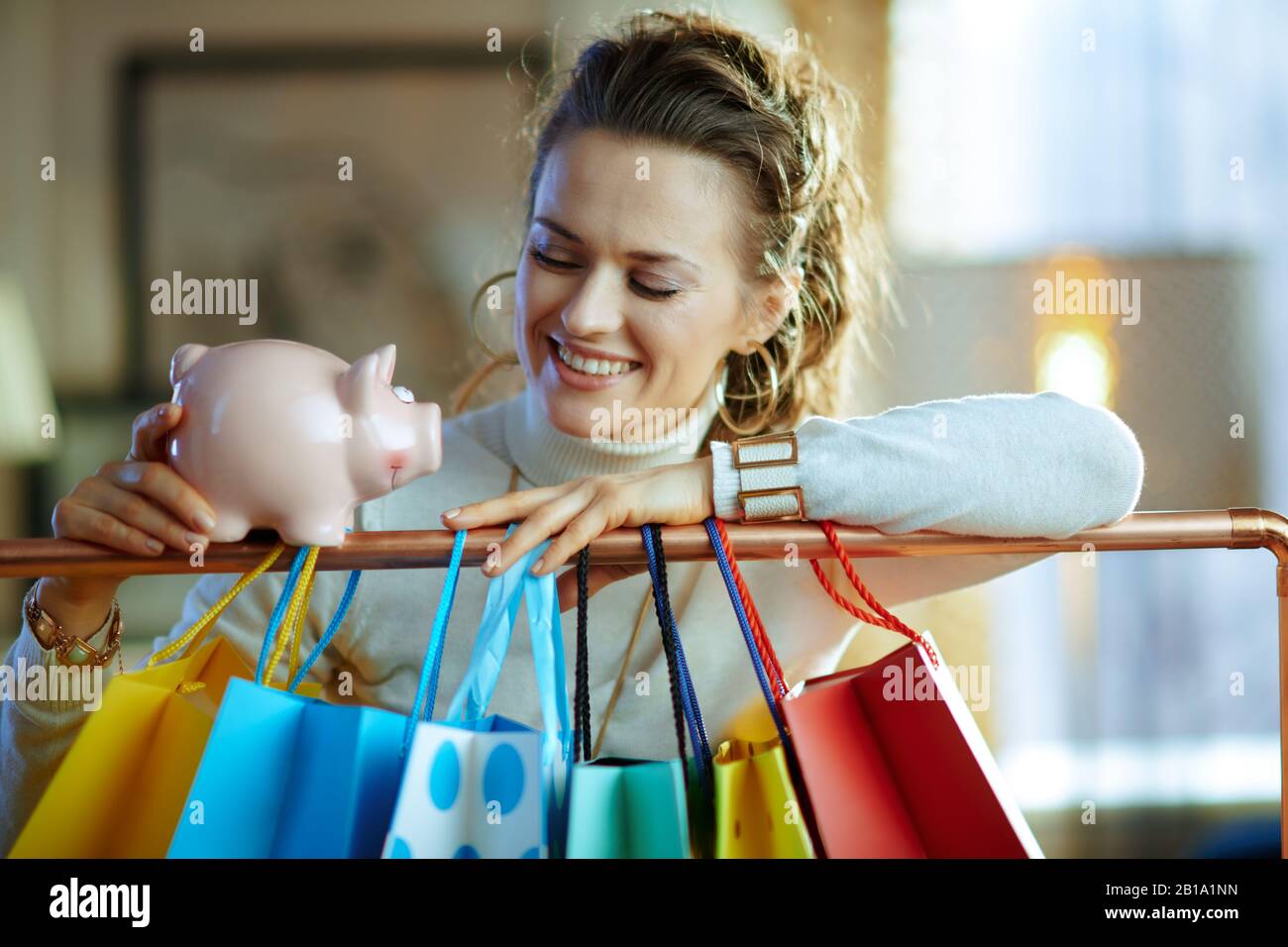 femme souriante d'âge moyen en sweat-shirt blanc et jupe près de sacs à provisions colorés accrochés sur rail de vêtements en cuivre avec une banque de porc. Banque D'Images